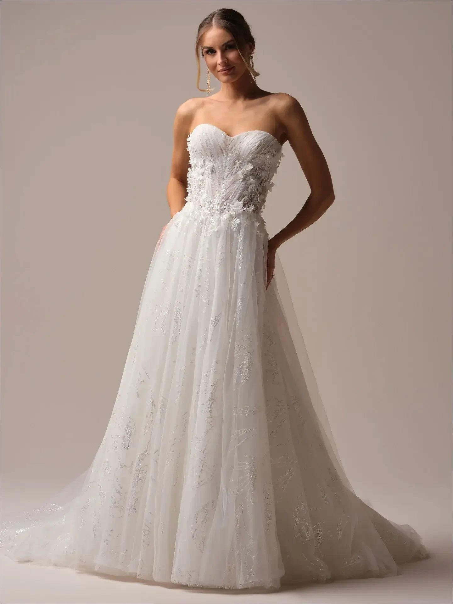 Model is wearing a strapless sparkly A-line wedding gown with a flowing tulle skirt and 3D floral appliqu&eacute;s on the bodice.