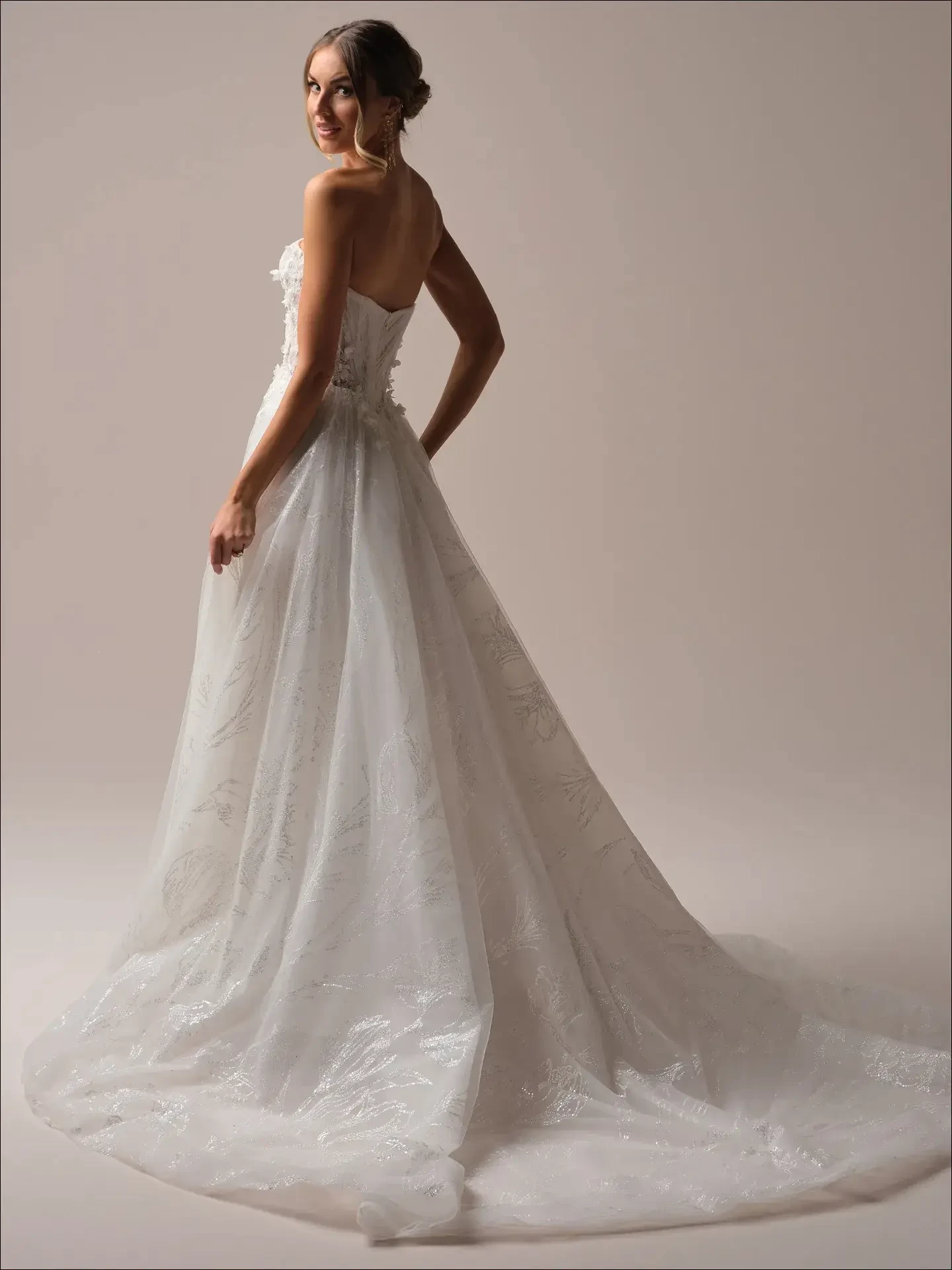 Model is wearing a strapless sparkly A-line wedding gown with a flowing tulle skirt and 3D floral appliqu&eacute;s on the bodice.